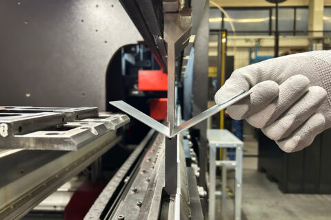 Dettaglio di una macchina dedicata alla piegatura lamiera in fase di piegatura di un pezzo di lamiera piuttosto piccolo sostenu da una mano di un operatore.